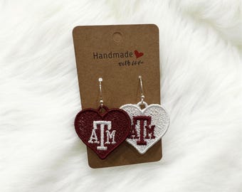 May include: Heart-shaped embroidered earrings, one maroon and one white, each featuring the letters "ATM" in a contrasting color. The earrings hang from silver-colored hooks and are displayed on a brown card that reads "Handmade with love."