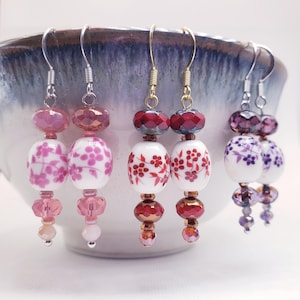 Może przedstawiać: Kolekcja pięciu par kolczyków wiszących. Każda para charakteryzuje się unikalną kombinacją koralików, w tym fasetowanych i okrągłych w odcieniach różu, czerwieni i fioletu. Kolczyki mają kwiatowe wzory i są zawieszone na srebrnych lub złotych haczykach.