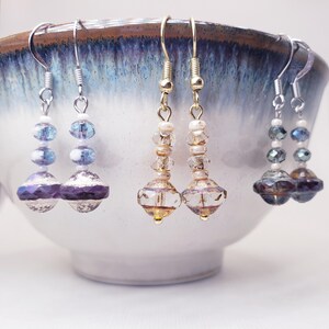 May include: Three pairs of dangle earrings featuring faceted glass beads in purple, clear, and dark blue tones. Each earring is suspended from a metal hook and accented with small white spacer beads, creating a tiered design with a reflective finish.