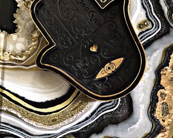 Könnte beinhalten: Eine schwarze Hamsa-Hand-Schale mit goldfarbenem Rand, verziert mit einem geprägten Augensymbol, einem kleinen Herz und verschlungenen Linienmustern. Die Schale liegt auf einer größeren Oberfläche im Geoden-Stil mit schwarzen, weißen und goldenen Akzenten.