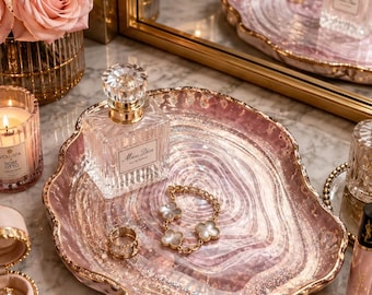 May include: An irregular, oval-shaped decorative tray featuring a translucent pink and white marbled resin surface with gold-toned glitter accents and a metallic gold-painted scalloped edge, holding a glass perfume bottle and jewellery.