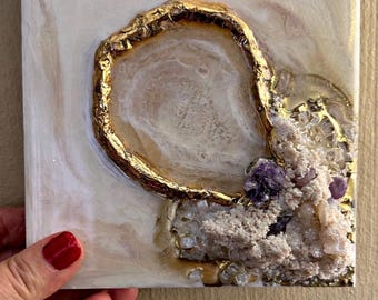 Könnte beinhalten: Quadratische Kunst mit einem goldumrandeten Geoden-Design und einer zentralen Öffnung. Das Kunstwerk verwendet Beige-, Weiß- und Goldtöne mit einem Cluster aus Kristallen und violetten Akzenten. Ein einzigartiger Dekorationsartikel.