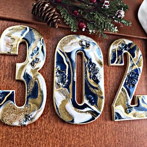 May include: Three resin house numbers, 302, in a blue, white and gold abstract design. The numbers are mounted on a wooden surface.