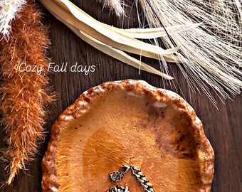 Könnte beinhalten: Eine runde, texturierte, bernsteinfarbene Keramikschale mit unebenem, gewelltem Rand. In der Schale liegt ein goldfarbenes Metallkettenarmband mit einem zentralen CD-Logo-Anhänger. Der Text "Cozy Fall days" ist links in Weiß aufgedruckt.