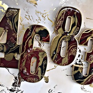 May include: Four resin numbers, 4, 6, 8, and 3, in a burgundy and gold colour scheme with gold glitter and red glitter accents. The numbers are in a resin art style with a marbled effect.