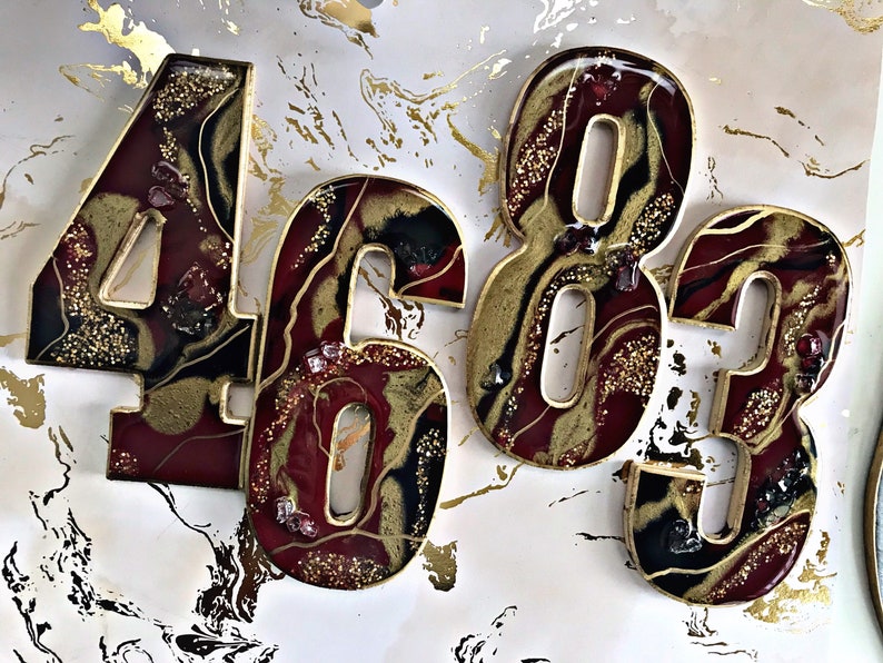 May include: Four resin numbers, 4, 6, 8, and 3, in a burgundy and gold colour scheme with gold glitter and red glitter accents. The numbers are in a resin art style with a marbled effect.