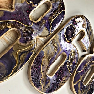 May include: Two resin number ornaments in the shape of the number 80. The ornaments are painted with a purple, gold, and white marbled design and have a glittery finish.