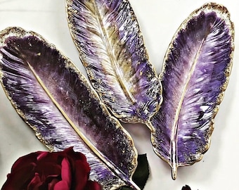 Könnte beinhalten: Drei federförmige Dekoschalen in tiefen Violett- und Weißtönen mit Blattgoldakzenten an den Rändern. Die Oberflächen haben ein glänzendes Finish mit einer sichtbaren Textur, die an natürliche Federadern und einen Kiel erinnert.