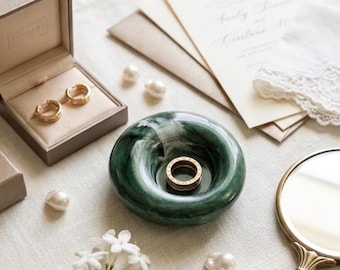 Könnte beinhalten: Eine kreisförmige Ringablage aus dunkelgrünem Marmor mit einem Goldring, platziert neben einer beigen Schmuckschatulle mit goldenen Creolen, mehreren weißen Perlen, einem kleinen Handspiegel mit goldenem Griff und einem Zweig weißer Blüten.