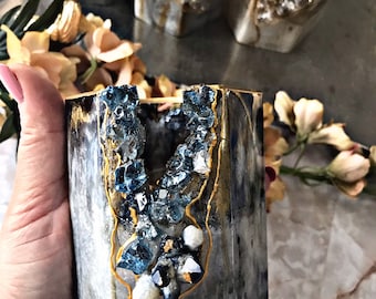 May include: A decorative, cylindrical container with a geode-like interior. The container has a marbled grey and black exterior with gold accents. The interior is filled with blue and clear crystal formations, creating a unique, artistic piece.