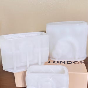 Può includere: Tre stampi in silicone bianco traslucido per colata, caratterizzati da forme rettangolari e arrotondate con motivi in rilievo. Gli stampi sono disposti su una scatola color pesca chiaro con la parola LONDON stampata in lettere maiuscole nere.