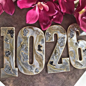 May include: Four resin house numbers, 1026, in a white, grey, and gold colour scheme with a glittery finish. The numbers have a marbled effect and are decorated with black and silver glitter.