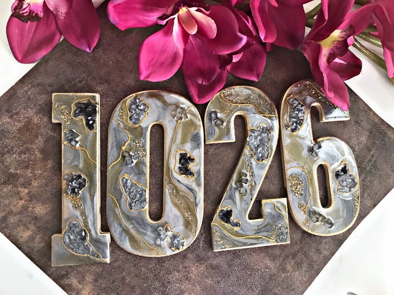 May include: Four resin house numbers, 1026, in a white, grey, and gold colour scheme with a glittery finish. The numbers have a marbled effect and are decorated with black and silver glitter.