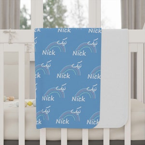 Könnte beinhalten: Eine blaue Babydecke mit weißem Hintergrund. Die Decke hat ein sich wiederholendes Muster aus Regenbögen und den Namen "Nick" in weiß.