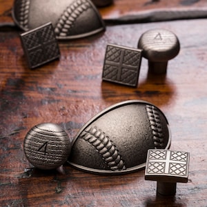 May include: A set of six metal cabinet knobs in a baseball theme. The knobs are in a distressed silver finish and feature a variety of baseball-inspired designs, including a baseball, a baseball glove, and a baseball diamond.