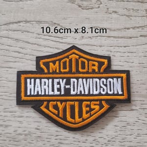 May include: Embroidered Harley-Davidson patch, measuring 10.6cm by 8.1cm. The patch features the Harley-Davidson logo in orange and white on a black background. The text "MOTOR HARLEY-DAVIDSON CYCLES" is prominently displayed.