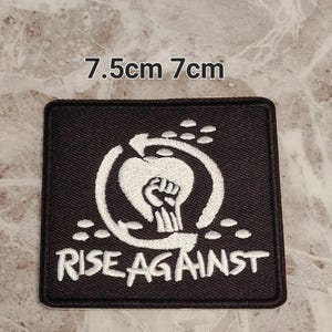 Può includere: Toppa quadrata nera con design e testo ricamati in bianco. Il design presenta un pugno chiuso all'interno di un cerchio a forma di cuore, con le parole "RISE AGAINST" sotto. La toppa misura 7,5 cm x 7 cm.
