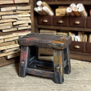 May include: A small, weathered wooden stool, painted in brown, black, red, and orange hues. The miniature stool has a square seat and four legs, with visible wood grain, giving it a rustic appearance. Ideal for a dollhouse or miniature setting.