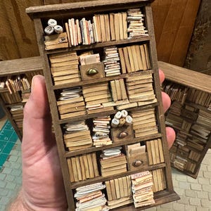 Może przedstawiać: Miniaturowa drewniana półka na książki wypełniona maleńkimi książkami, zwiniętymi papierami i małymi szufladami. Półka na książki jest w kolorze ciemnobrązowym i ma wiele półek. Książki mają postarzone, pożółkłe strony, a ogólna estetyka jest vintage.