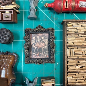 Puede incluir: Una colección de decoración en miniatura de temática gótica, que incluye un cuadro enmarcado de esqueletos, una estatua de gárgola, un buzón rojo, una estantería en miniatura y un sillón marrón. Los artículos están dispuestos sobre una esterilla de corte verde.