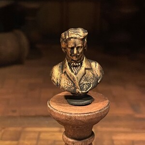 May include: A small, antique-style bust of a man with a mustache and a jacket, mounted on a wooden pedestal. The bust has a bronze finish with dark accents, and the pedestal is a warm brown colour.