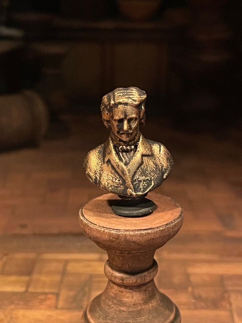 May include: A small, antique-style bust of a man with a mustache and a jacket, mounted on a wooden pedestal. The bust has a bronze finish with dark accents, and the pedestal is a warm brown colour.