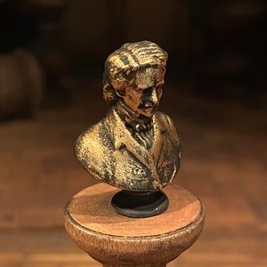 May include: A small, bronze-effect bust of a man with long hair and a jacket, mounted on a wooden pedestal. The bust has a detailed face and is likely a decorative item or collectible.
