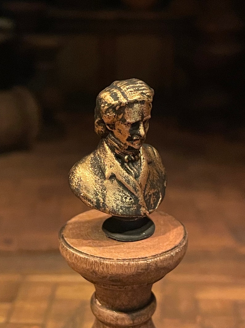 May include: A small, bronze-effect bust of a man with long hair and a jacket, mounted on a wooden pedestal. The bust has a detailed face and is likely a decorative item or collectible.