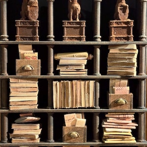 Op de afbeelding: Een donkerbruine houten boekenplank met gewelfde bovenste secties. De bovenste plank bevat drie kleine beelden. De onderste planken tonen stapels oude boeken, papieren en kleine houten lades met messing handgrepen.