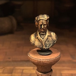 May include: A bronze-coloured bust of a man with detailed facial features and hair, displayed on a small wooden pedestal. The bust is the central element, highlighting the intricate details of the sculpture. The pedestal is round and has a dark finish.