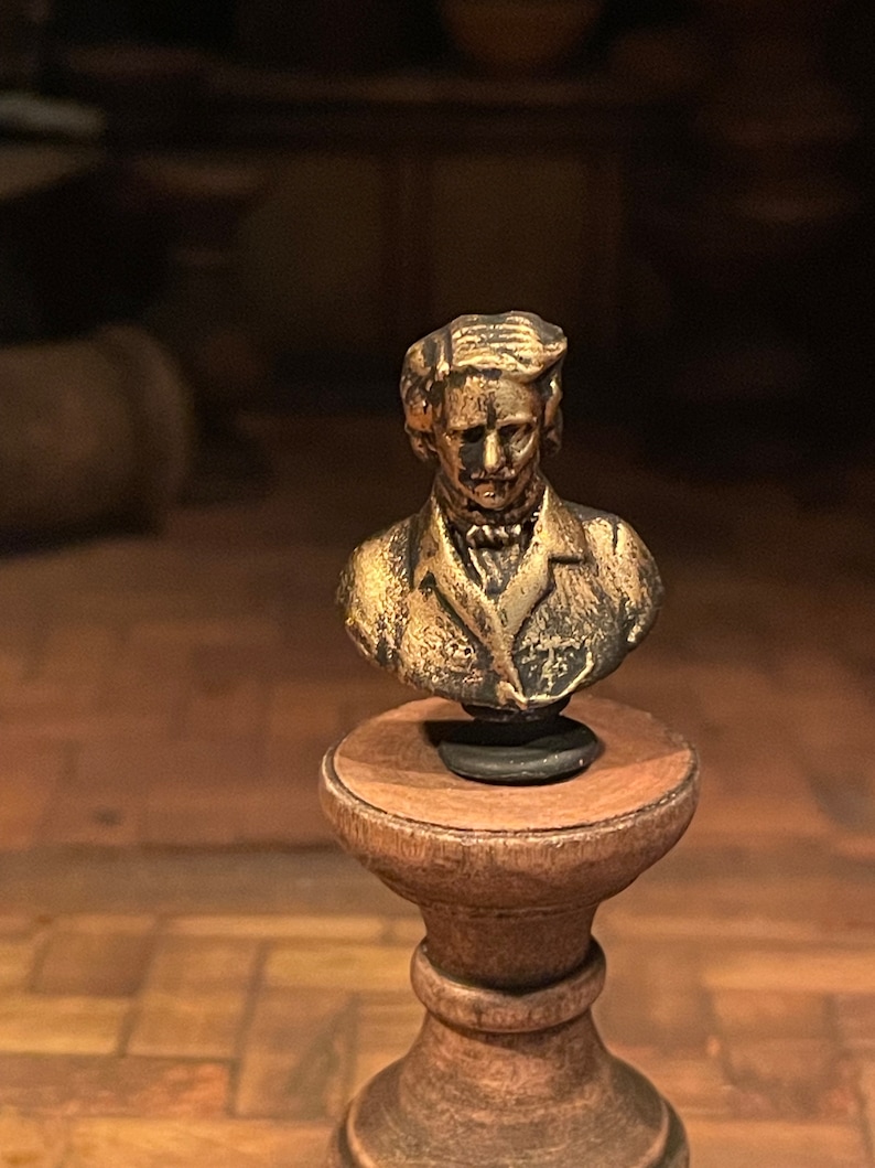 May include: A bronze-coloured bust of a man with detailed facial features and hair, displayed on a small wooden pedestal. The bust is the central element, highlighting the intricate details of the sculpture. The pedestal is round and has a dark finish.
