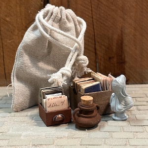 Op de afbeelding: Een miniatuurset met een beige jute zakje met trekkoord, twee kleine bruine opbergdoosjes gevuld met kleine gestapelde papieren en boeken, een kleine bruine kleipot met een kurken stop en een lichtgrijs beeldje van een persoon met hoofdbedekking.