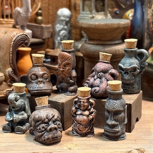 May include: A selection of small, brown decorative bottles with cork stoppers. The bottles are sculpted in various shapes, including a monkey, a tiki, an octopus, a skull, and a Buddha. The bottles are arranged on a wooden surface.
