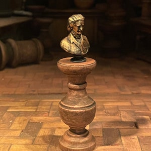 May include: A miniature gold-coloured bust of a man on a wooden pedestal. The bust has detailed facial features and sits on a small, round black base. The pedestal is light brown and has a decorative, turned design.