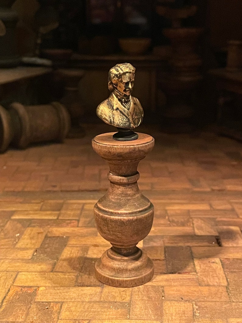 May include: A miniature gold-coloured bust of a man on a wooden pedestal. The bust has detailed facial features and sits on a small, round black base. The pedestal is light brown and has a decorative, turned design.