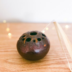 May include: A dark brown ceramic sphere with multiple holes, possibly a diffuser or decorative object. The sphere sits on a wooden surface, with a dried leaf and string attached. The object has a glossy finish and a few orange paint marks.