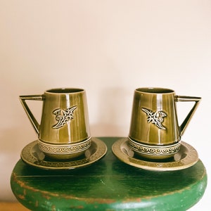 May include: Two olive green ceramic mugs with matching saucers. Each mug features a geometric handle and a decorative emblem. The saucers have a detailed border design. The mugs and saucers are displayed on a green stool.