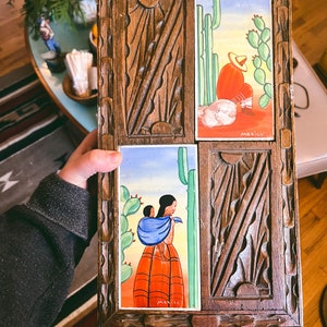 May include: A wooden decorative box with four ceramic tiles featuring Mexican folk art. The tiles depict scenes of people and cacti. The box has a carved wooden frame with a sunburst design. The word "Mexico" is visible on the tiles.