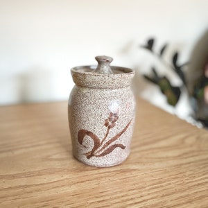 May include: A speckled ceramic jar with a lid. The jar is beige with brown floral designs. The jar is sitting on a wooden surface.