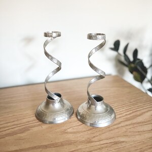 May include: Two silver candlesticks with a hammered texture base and a spiral stem. The candlesticks are displayed on a wooden surface. The design is modern and elegant, suitable for home decor.