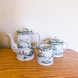 May include: A vintage porcelain tea set featuring a teapot, creamer, and sugar bowl. The set is decorated with a landscape scene in shades of blue, green, and white, with a green and floral border. The teapot has a curved spout and handle.