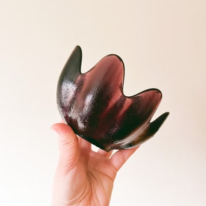 May include: A dark purple glass bowl with a unique, petal-like shape. The bowl has a glossy finish and is held up by a hand. The bowl's design is reminiscent of a flower or leaf, adding a touch of elegance.