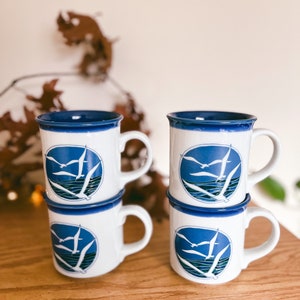 May include: Four white ceramic mugs with a blue rim and handle are stacked on a wooden surface. Each mug features a blue circular design with seagulls flying over water. The mugs are a coastal theme.