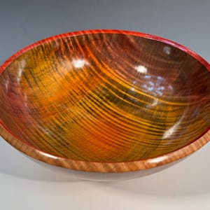 May include: A large wooden bowl with a red rim. The bowl is made of wood with a beautiful grain pattern in shades of orange, yellow, and brown.