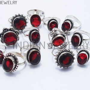 Può includere: Una collezione di 12 anelli d'argento con pietre preziose rosse. Gli anelli hanno disegni intricati e sono disposti su una superficie bianca.