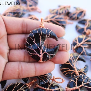 Könnte beinhalten: Schwarzer Obsidian-Halbmond-Anhänger mit einem Baumdesign aus Kupferdraht. Der Anhänger wird in einer Hand gehalten.