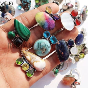 Op de afbeelding: Een verzameling zilveren ringen met verschillende gekleurde edelstenen, waaronder turquoise, malachiet, amethist en regenboog-maansteen. De ringen worden getoond op een hand, waardoor de ingewikkelde details en unieke ontwerpen worden getoond.