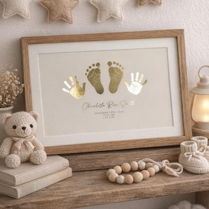 Puede incluir: Una impresión enmarcada con huellas de manos y pies doradas, el nombre "Charlotte Rose Smith" y detalles del nacimiento. Un pequeño oso de peluche de ganchillo y patucos para bebé están en una estantería de madera. Estrellas decorativas en el fondo.