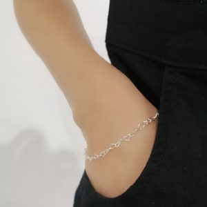May include: A delicate silver bracelet featuring a chain of small heart-shaped links. The bracelet is worn on a wrist, partially visible against a black garment. The overall aesthetic is minimalist and elegant, suitable for everyday wear.