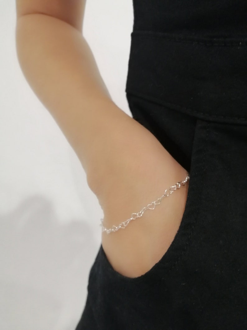May include: A delicate silver bracelet featuring a chain of small heart-shaped links. The bracelet is worn on a wrist, partially visible against a black garment. The overall aesthetic is minimalist and elegant, suitable for everyday wear.